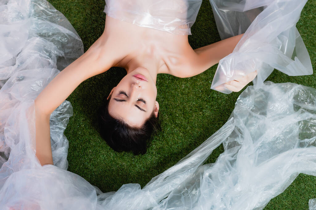 Draufsicht der jungen brünetten Frau in Polyethylen gehüllt auf Gras liegend, ökologisches Konzept  - Foto, Bild