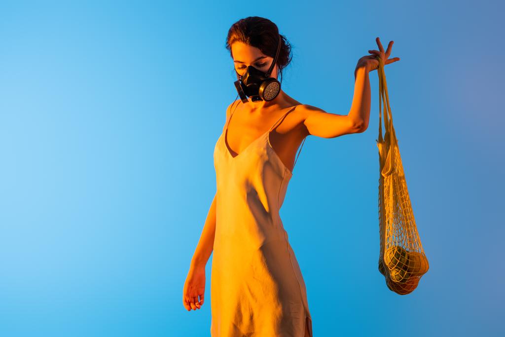 joven morena mujer en máscara de gas y vestido de seda sosteniendo bolsa de hilo reutilizable con frutas en azul - Foto, Imagen