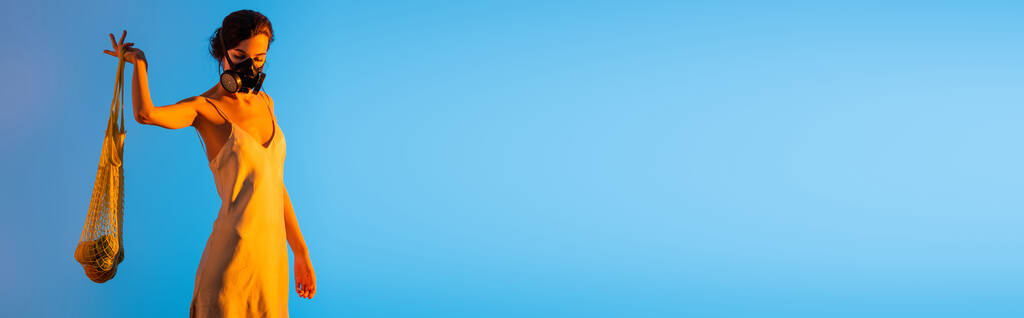 cultivo panorámico de mujer en máscara de gas y vestido de seda sosteniendo bolsa de hilo reutilizable con frutas en azul - Foto, imagen
