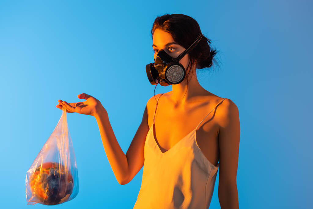 γυναίκα σε μάσκα αερίου κρατώντας πλαστική σακούλα με σφαίρα σε μπλε, έννοια οικολογία  - Φωτογραφία, εικόνα