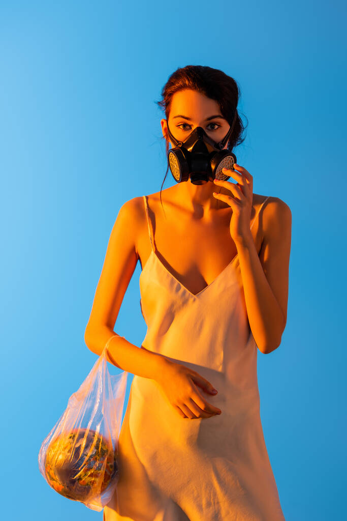 kobieta dotykająca maski gazowej i trzymająca plastikową torbę z kulą na niebieskiej, ekologicznej koncepcji  - Zdjęcie, obraz