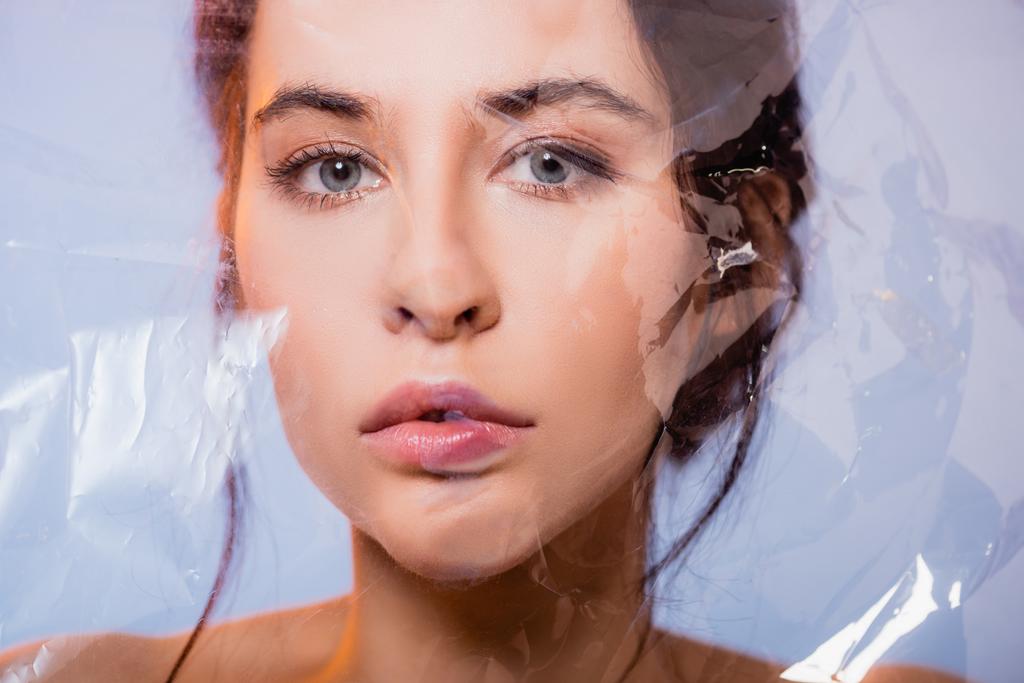 brunette jonge vrouw kijken naar camera door polyethyleen op grijs, ecologie concept  - Foto, afbeelding