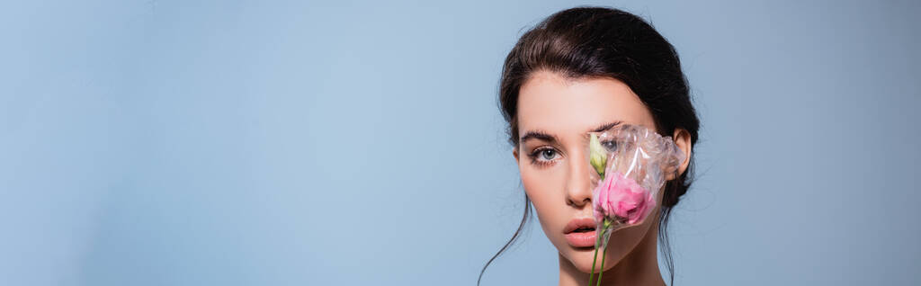 Panoramaaufnahme einer Frau, die das Auge mit Blumen aus Polyethylen bedeckt, isoliert auf blau, ökologisches Konzept  - Foto, Bild