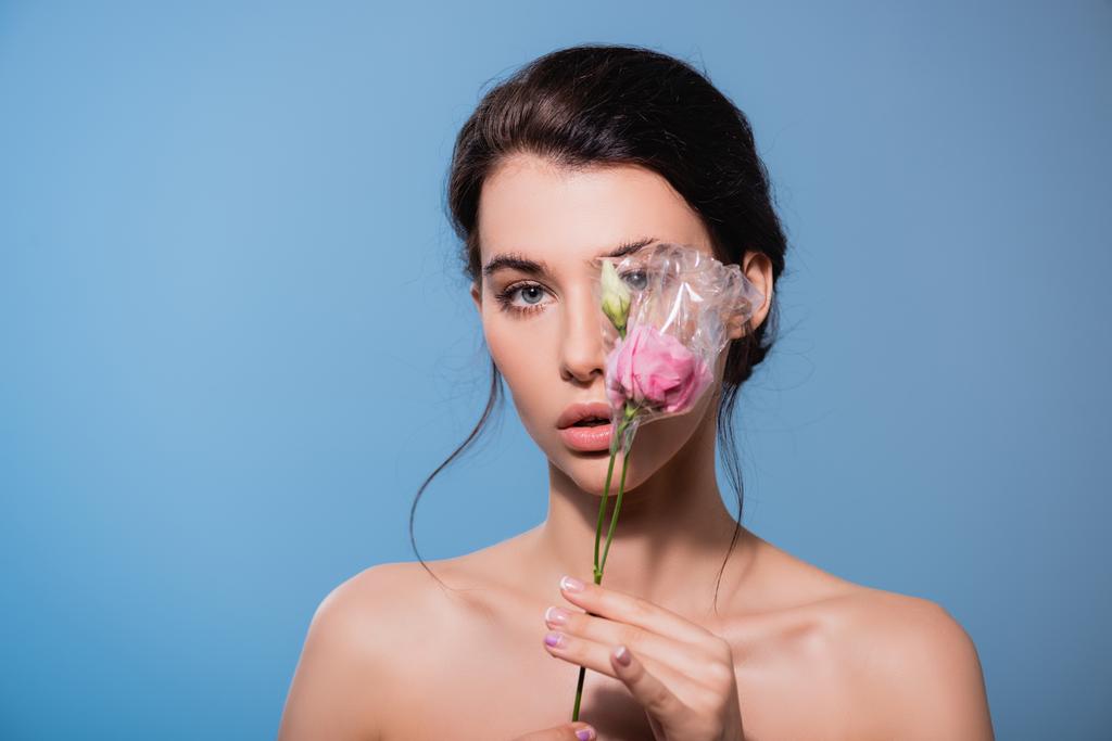 Genç ve çıplak bir kadın, mavi ve ekoloji kavramında izole edilmiş polietilen çiçeklerle gözlerini kapatıyor.  - Fotoğraf, Görsel