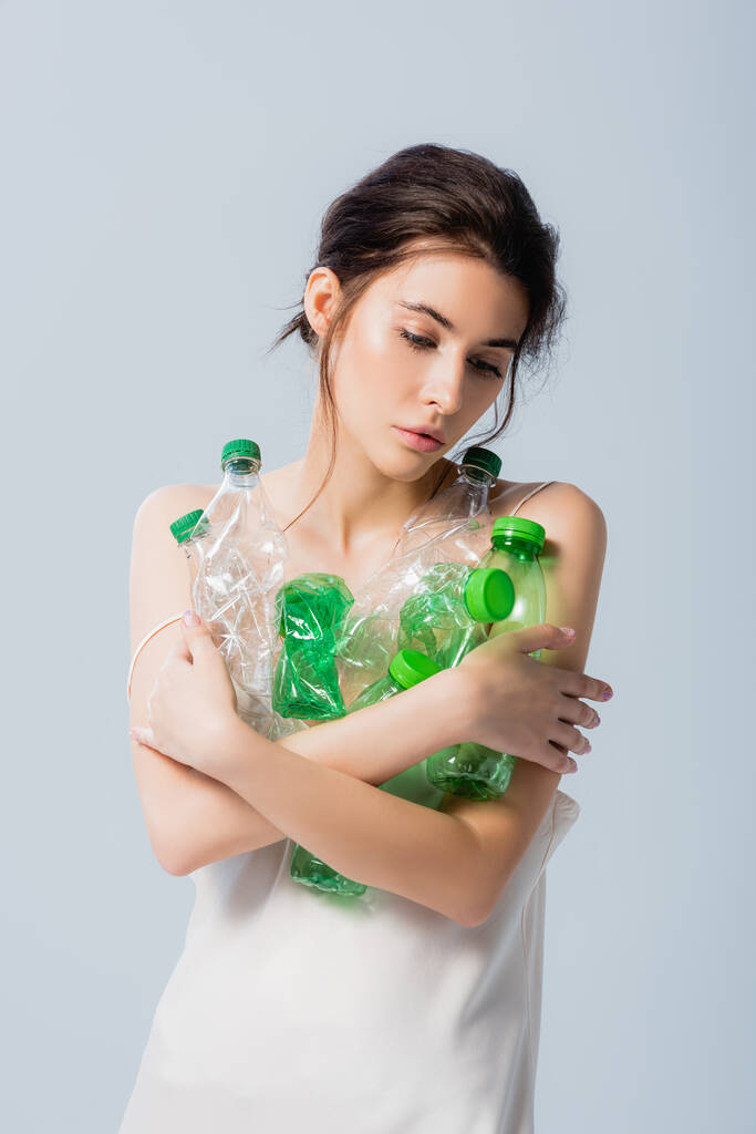 Esmer, ipek elbiseli, elinde plastik şişeler olan bir kadın. Gri, ekoloji kavramından soyutlanmış bir şekilde aşağıya bakıyor.  - Fotoğraf, Görsel