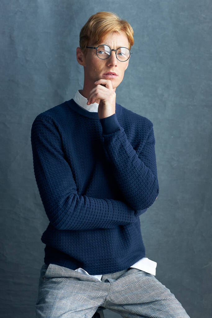 retrato de un joven pelirrojo con pecas sobre un fondo gris sentado en una silla con un suéter clásico azul oscuro en una camisa blanca, pantalones a cuadros grises, mira a la cámara a través de gafas transparentes  - Foto, imagen
