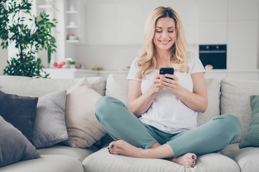 Photo of domestic pretty cheerful blond lady relaxing sit couch browsing telephone chatting friends enjoy quarantine stay home good mood legs crossed living room indoors - Photo, Image