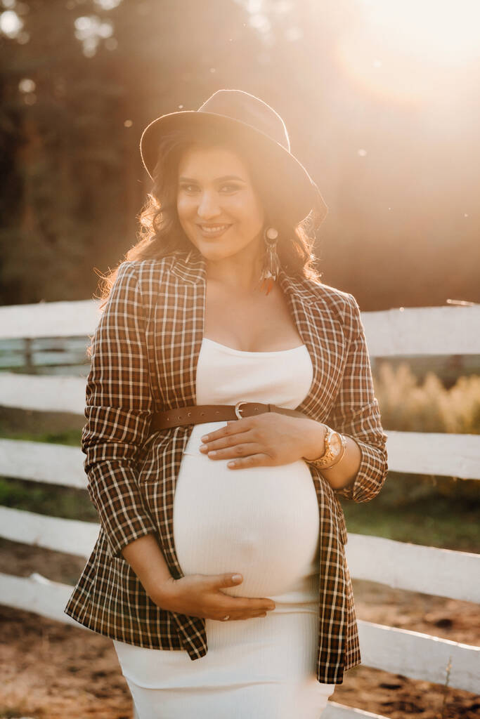 Gün batımında at ağılının yanında şapkalı hamile bir kız. Atlı kahverengi elbiseli şık hamile kadın. - Fotoğraf, Görsel