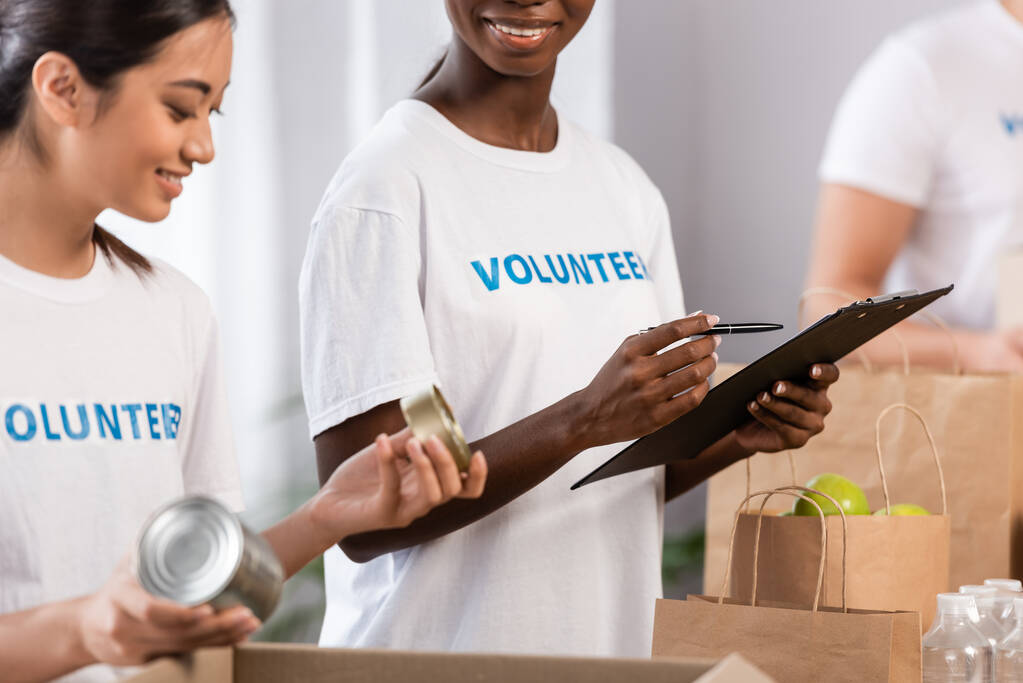 Selective focus of african american volunteer holding pen and clipboard near asian woman with tin cans and packages in charity center  - Photo, Image