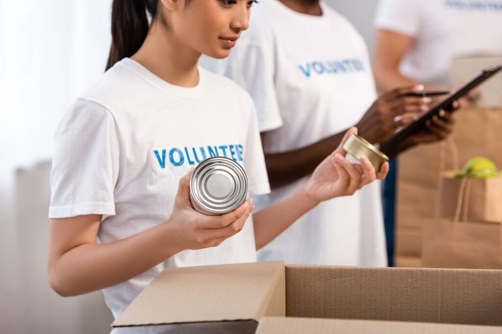 Selective focus of asian volunteer holding tin cans near cardboard box in charity center  - Photo, Image