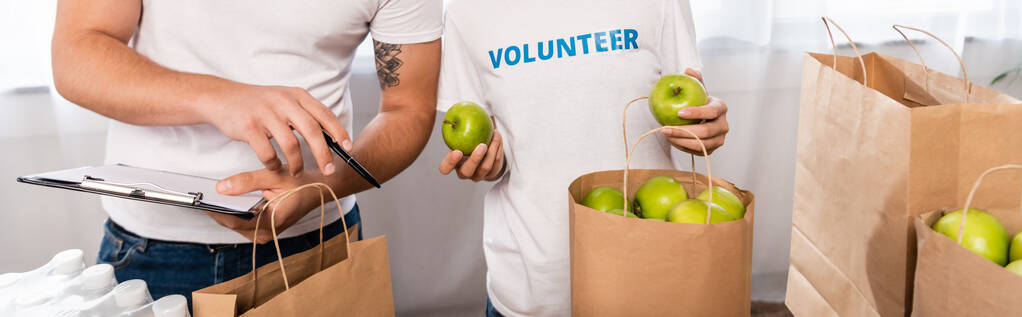 Récolte panoramique de bénévoles avec presse-papiers pointant à la main près d'une femme tenant des pommes et des paquets dans un centre caritatif  - Photo, image
