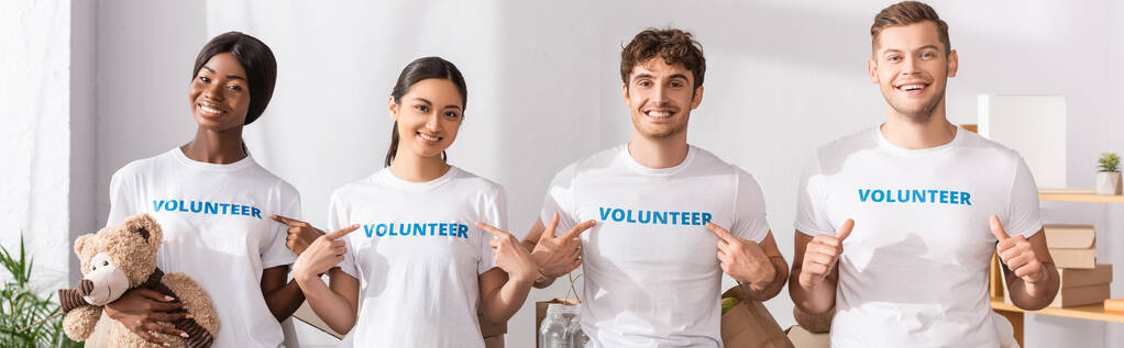 Horizontale oogst van jonge multi-etnische vrijwilligers wijzend met de vingers naar belettering op t-shirts en duimen laten zien in het liefdadigheidscentrum  - Foto, afbeelding