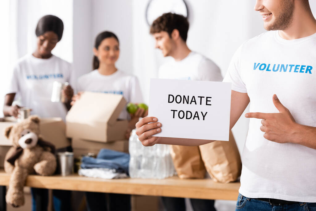 Selective focus of young volunteer pointing with finger at card with donate today lettering in charity center  - Photo, Image