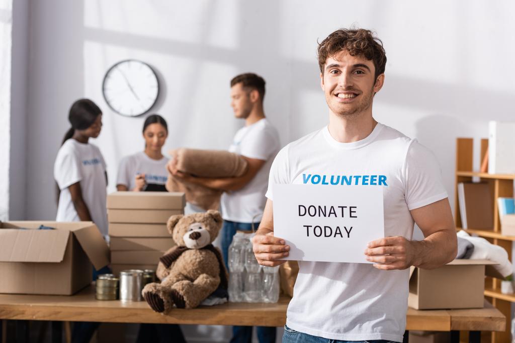 Селективный фокус волонтера с пожертвованием сегодня надпись на карточке глядя в камеру рядом с многонациональными людьми в благотворительном центре  - Фото, изображение