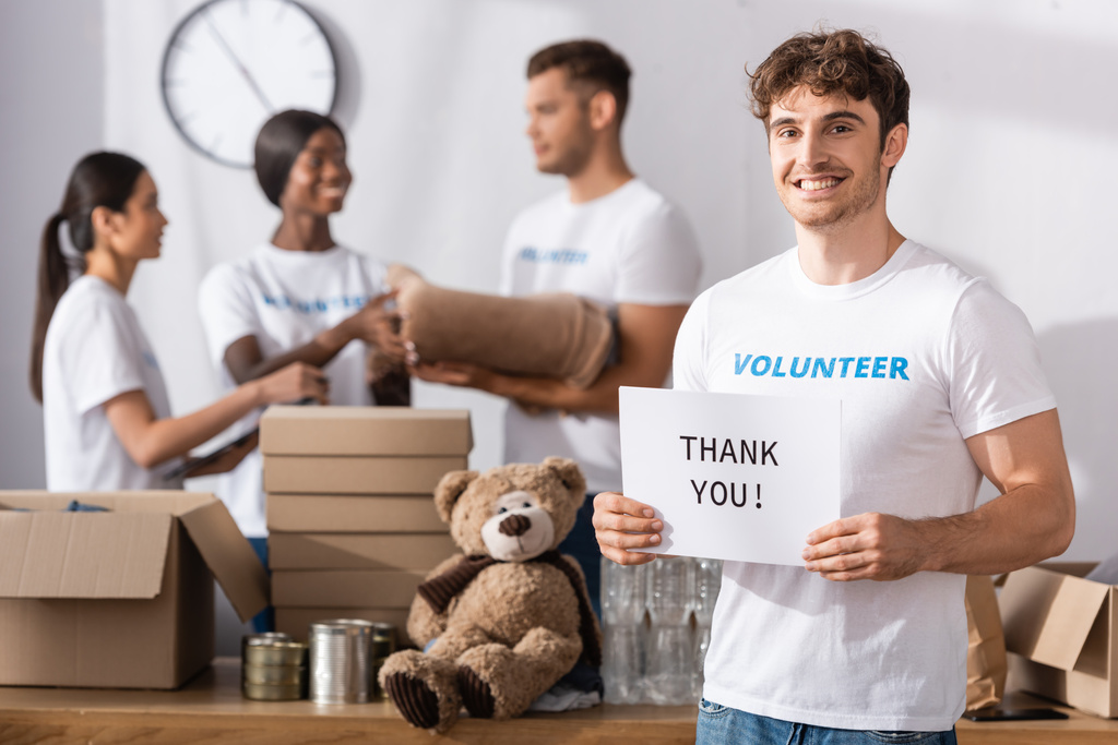 Selective focus of young volunteer holding card with thank you lettering with multicultural people at background in charity center  - Photo, Image
