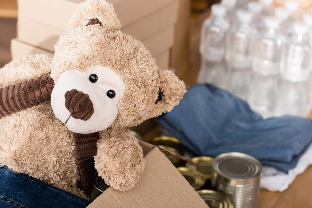 Селективный фокус мягкой игрушки в коробке возле жестяных банок и пожертвования в благотворительном центре  - Фото, изображение