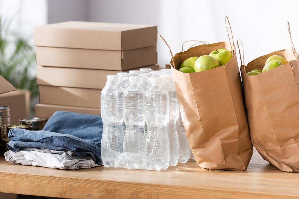 Sacchetti di carta con mele, bottiglie d'acqua e vestiti in tavola nel centro di beneficenza  - Foto, immagini