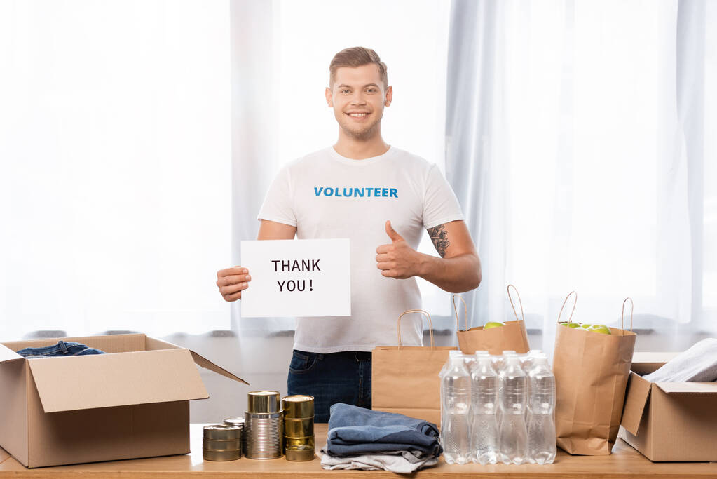 Εθελοντής που δείχνει όπως κρατώντας κάρτα με ευχαριστήρια γράμματα κοντά σε δωρεές στο τραπέζι  - Φωτογραφία, εικόνα