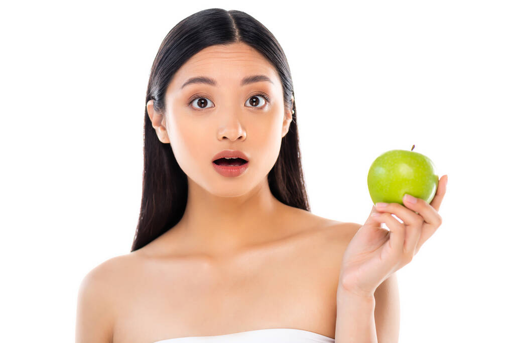 Удивлённая молодая азиатка смотрит в камеру и держит яблоко изолированным на белом - Фото, изображение