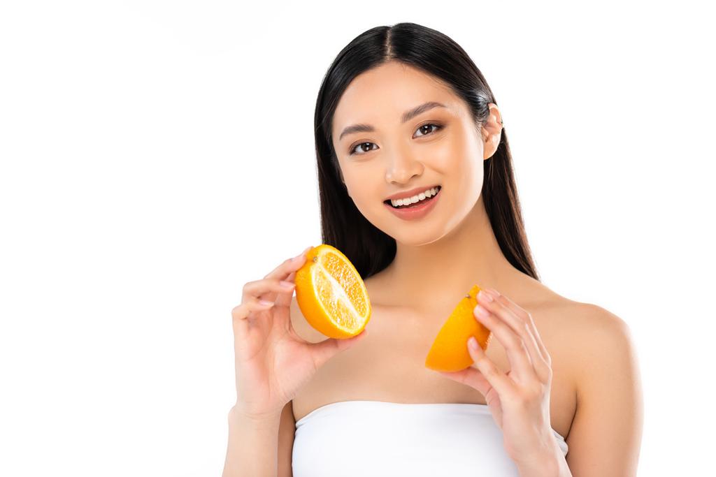 morena mujer asiática mirando a cámara mientras sostiene mitades de jugoso naranja aislado en blanco - Foto, Imagen