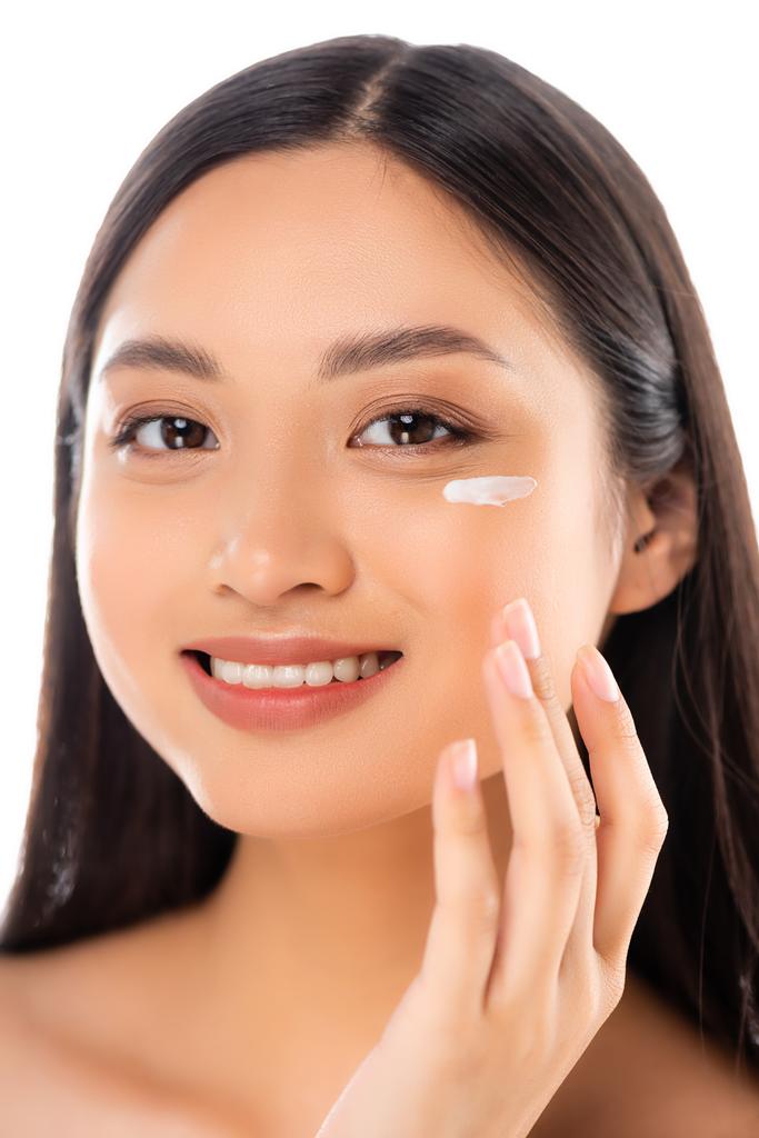 brünette asiatische Frau, die in die Kamera schaut, während sie kosmetische Creme auf das Gesicht aufträgt, isoliert auf weiß - Foto, Bild