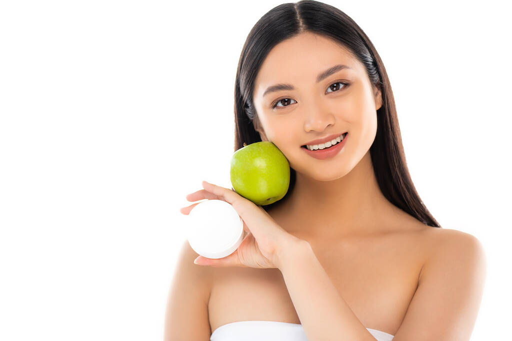 brunette asian woman holding green apple and cosmetic cream near face isolated on white - Photo, Image