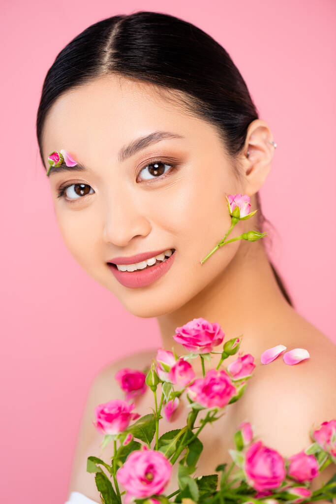 brunette aziatische vrouw met knoppen en bloemblaadjes op gezicht en schouder, met kleine rozen geïsoleerd op roze - Foto, afbeelding