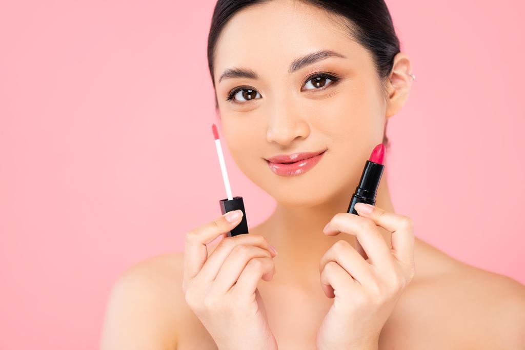 naked asian woman holding lipstick and lip gloss while looking at camera isolated on pink - Photo, Image