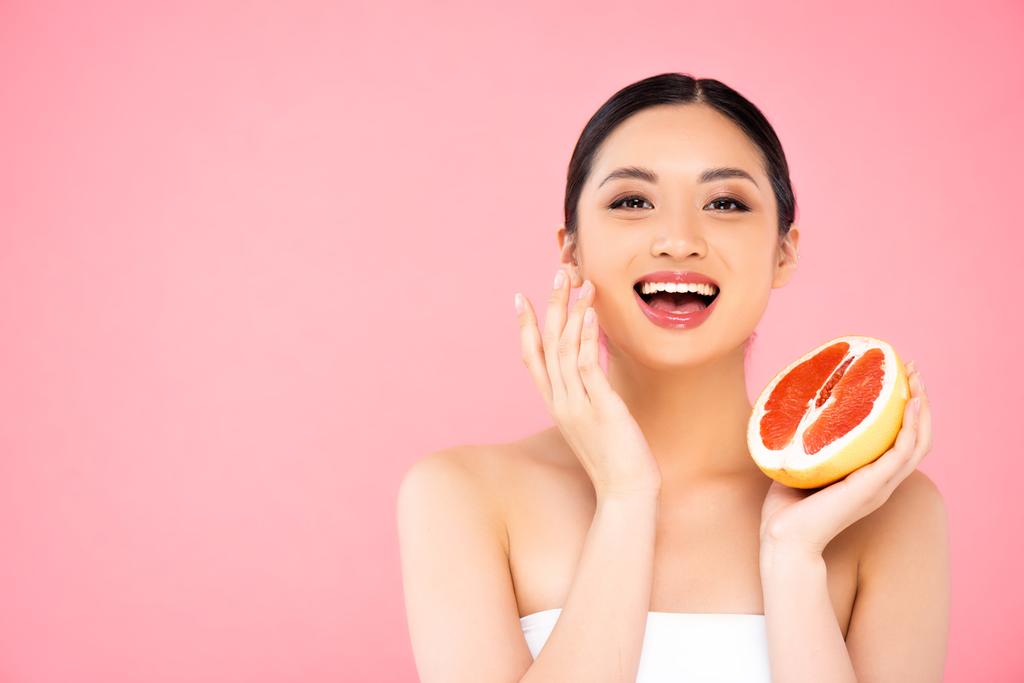 aufgeregte asiatische Frau hält die Hälfte der saftigen Grapefruit, während sie Gesicht isoliert auf rosa berührt - Foto, Bild