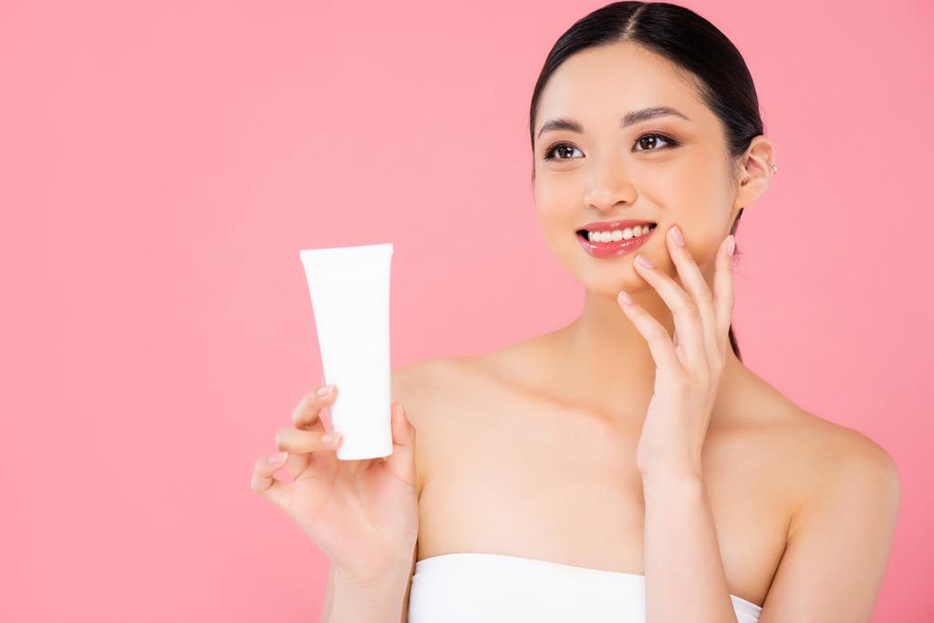 brunette asiatique femme tenant tube de crème cosmétique, toucher le visage et regarder loin isolé sur rose - Photo, image
