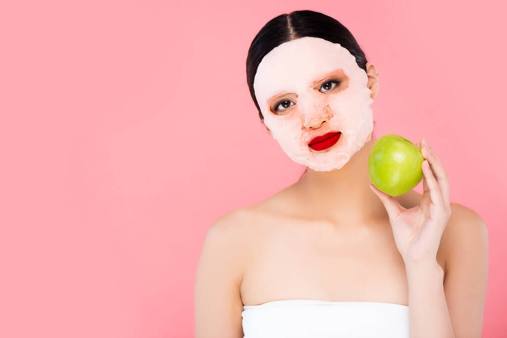 ázsiai nő arc maszk gazdaság zöld alma, miközben néz kamera elszigetelt rózsaszín - Fotó, kép