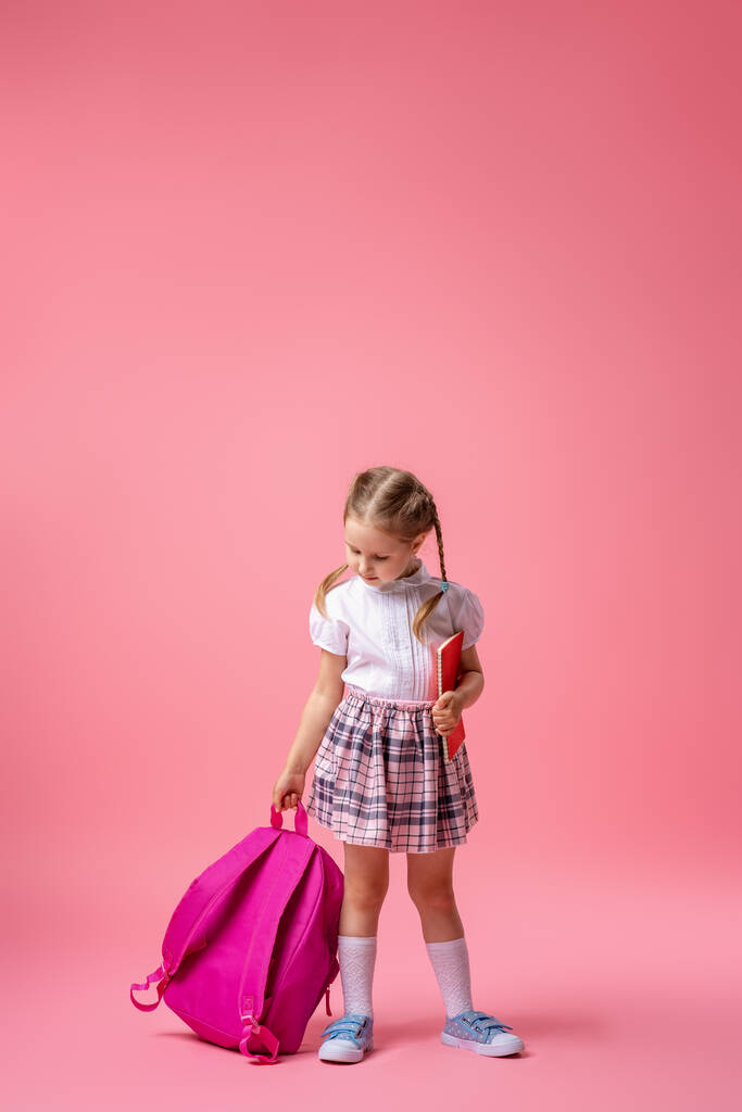 Glücklich lächelndes kleines Mädchen in Uniform mit Rucksack und Notizbuch in der Hand posiert im Studio auf rosa Hintergrund. Das Kind geht zum ersten Mal zur Schule. Zurück zur Schule. Kopierraum - Foto, Bild