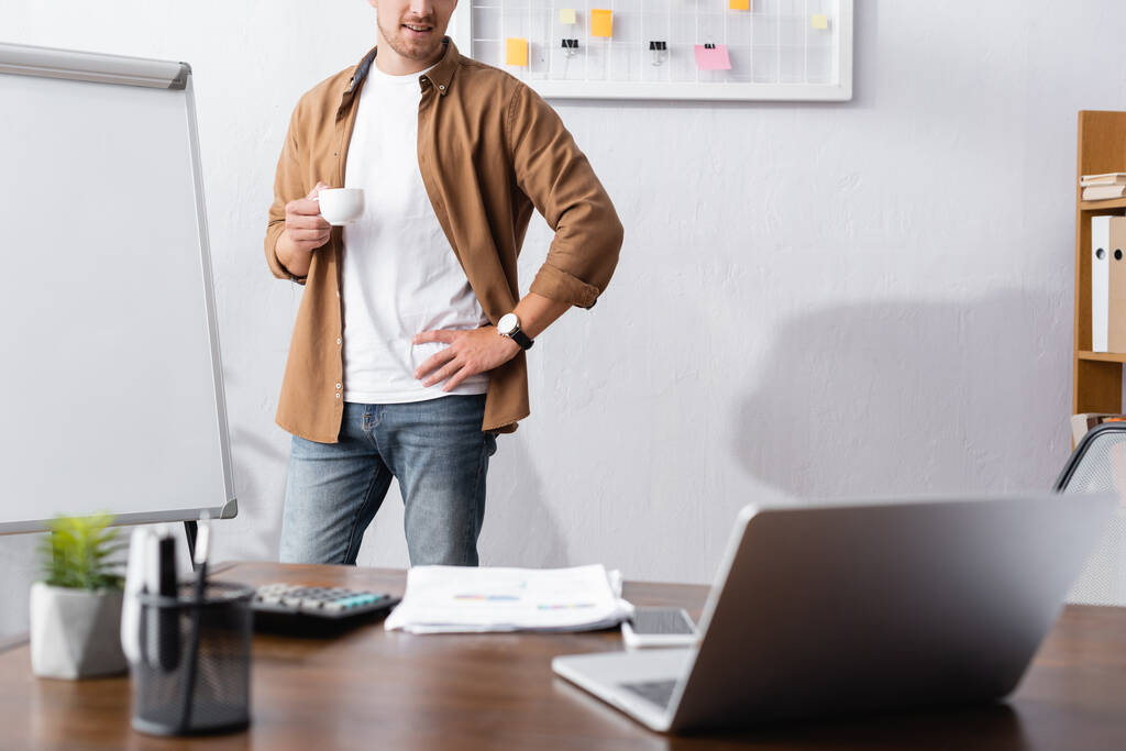 обрезанный взгляд бизнесмена в повседневной одежде держа чашку кофе, стоя с рукой на бедре в офисе - Фото, изображение