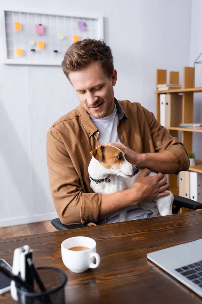üzletember alkalmi ruhákban ölelés Jack Russell terrier kutya, miközben ül a munkahelyen mellett egy csésze kávét - Fotó, kép
