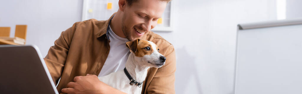 cosecha horizontal de joven empresario sosteniendo gato blanco russell terrier perro con manchas marrones en la cabeza en la oficina - Foto, imagen