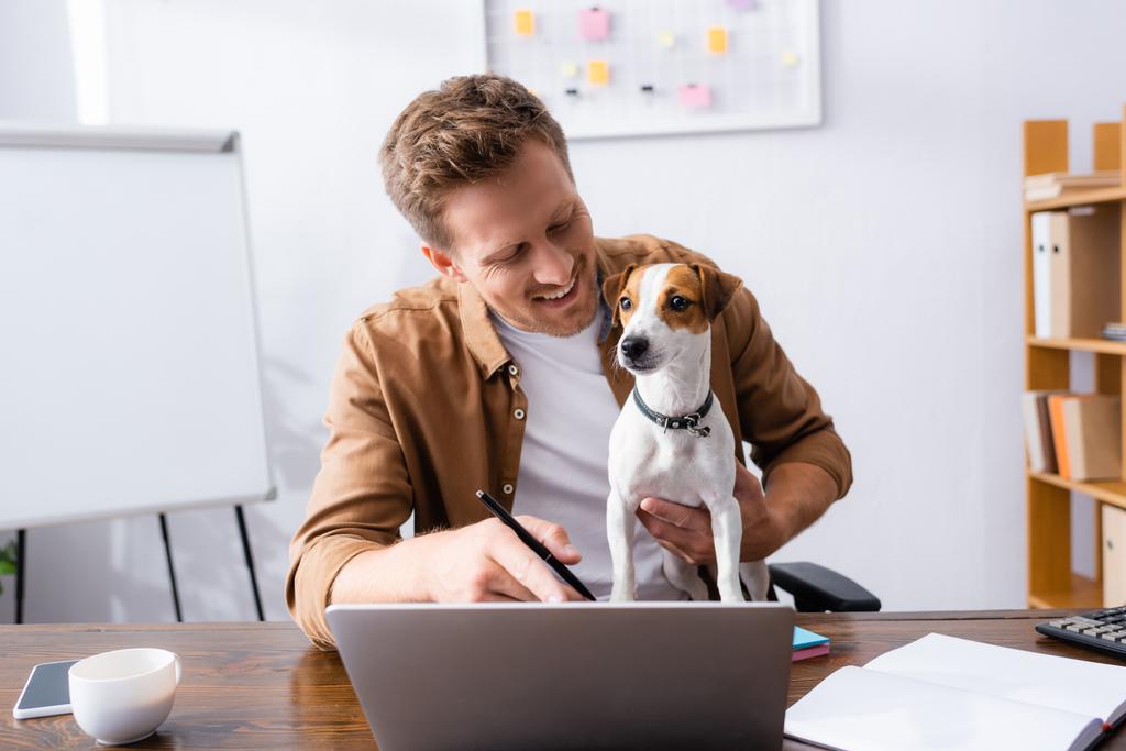 junger Geschäftsmann zeigt mit Finger auf Laptop, während er mit Jack Russell Terrier-Hund im Büro arbeitet - Foto, Bild
