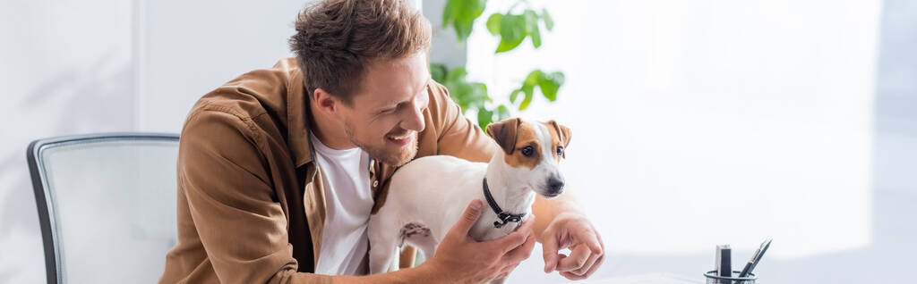 plano panorámico de joven hombre de negocios señalando con el dedo cerca de jack russell terrier perro en la oficina - Foto, Imagen
