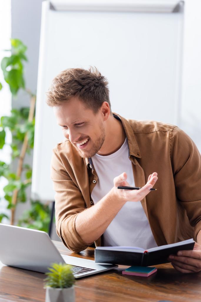 fiatal üzletember kezében notebook és toll közben video chat a laptopon - Fotó, kép