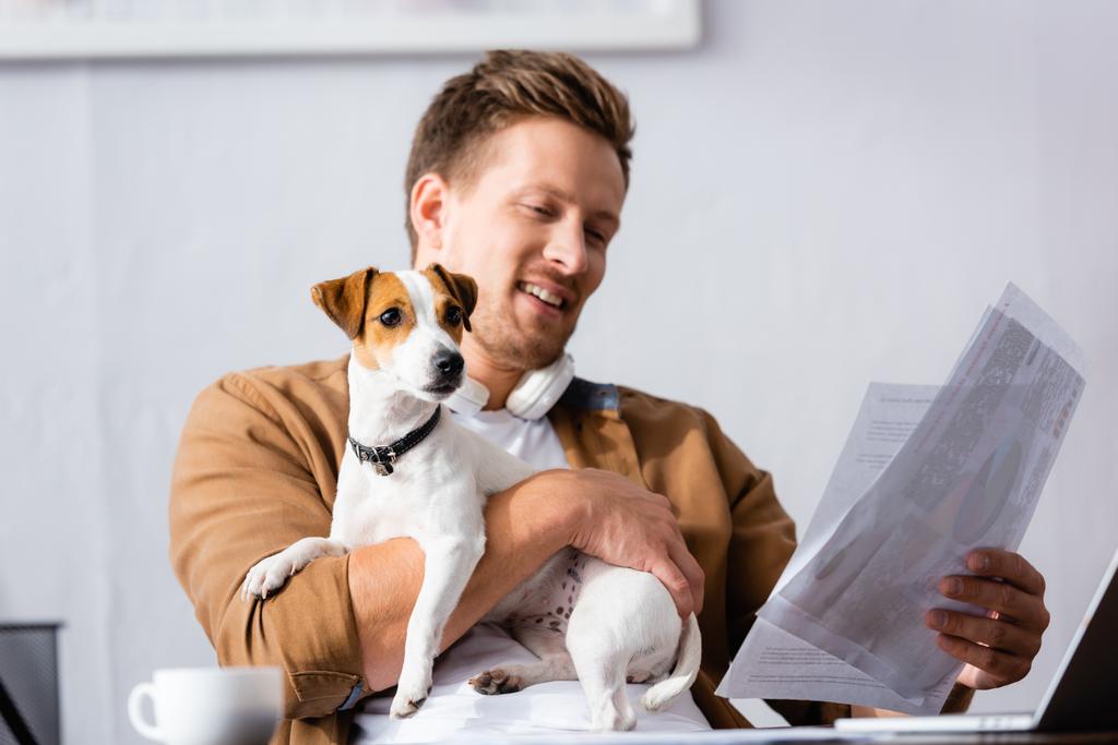 Jungunternehmer schaut sich Dokumente an, während er Jack Russell Terrier Dog am Arbeitsplatz knuddelt - Foto, Bild