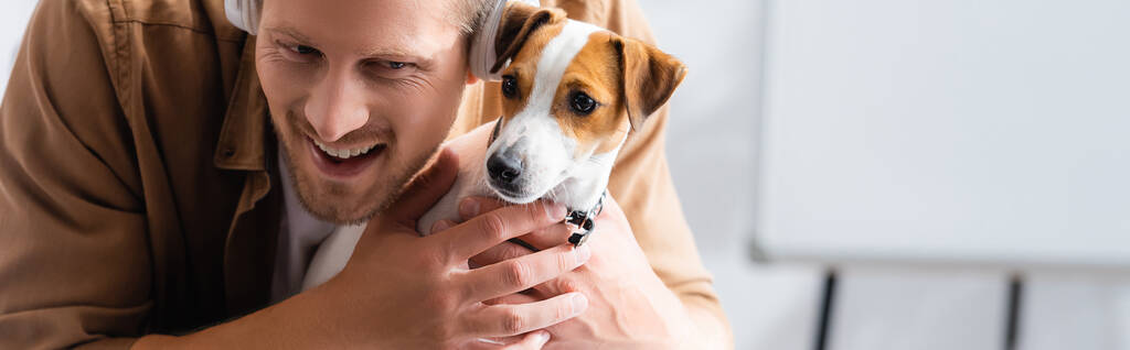 horizontales Bild eines Geschäftsmannes mit drahtlosen Kopfhörern, der Jack Russell Terrier Dog im Büro kuschelt - Foto, Bild