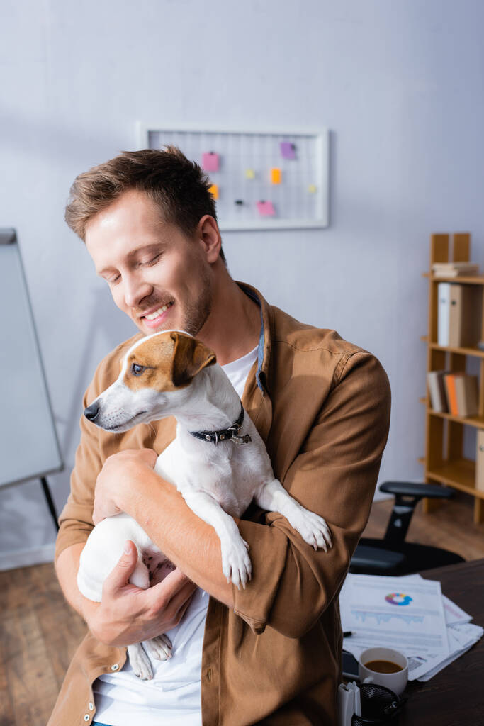 jeune homme d'affaires assis sur le bureau et tenant Jack Russell Terrier chien - Photo, image
