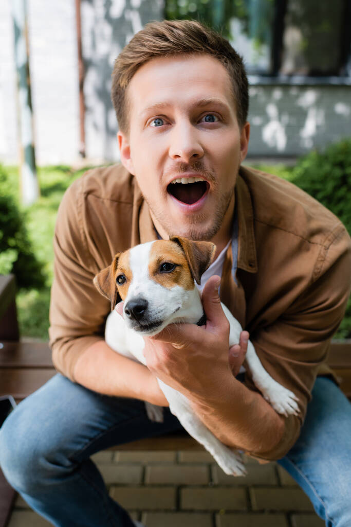 ενθουσιασμένος άνθρωπος με ανοιχτό στόμα κοιτάζοντας κάμερα, ενώ κρατώντας Jack Russell Terrier σκυλί - Φωτογραφία, εικόνα