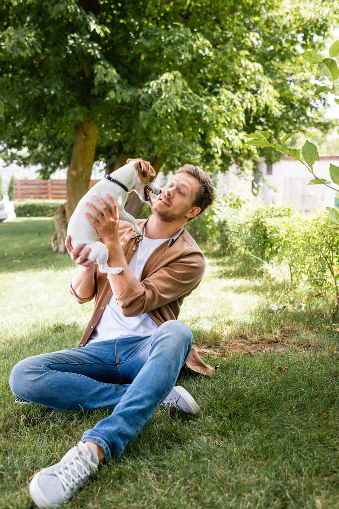 young man in casual clothes holding jack russell terrier dog while sitting on green grass - Photo, Image