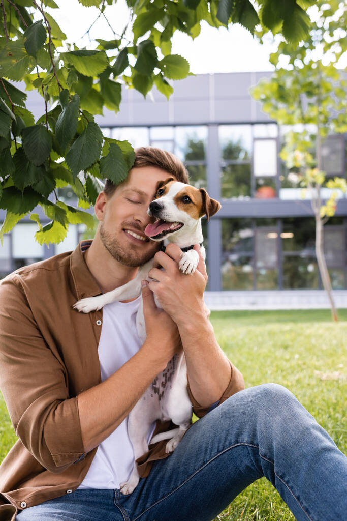νεαρός άνδρας με καφέ πουκάμισο και τζιν κάθεται στο γκαζόν και αγκαλιάζει Jack Russell Terrier σκυλί - Φωτογραφία, εικόνα