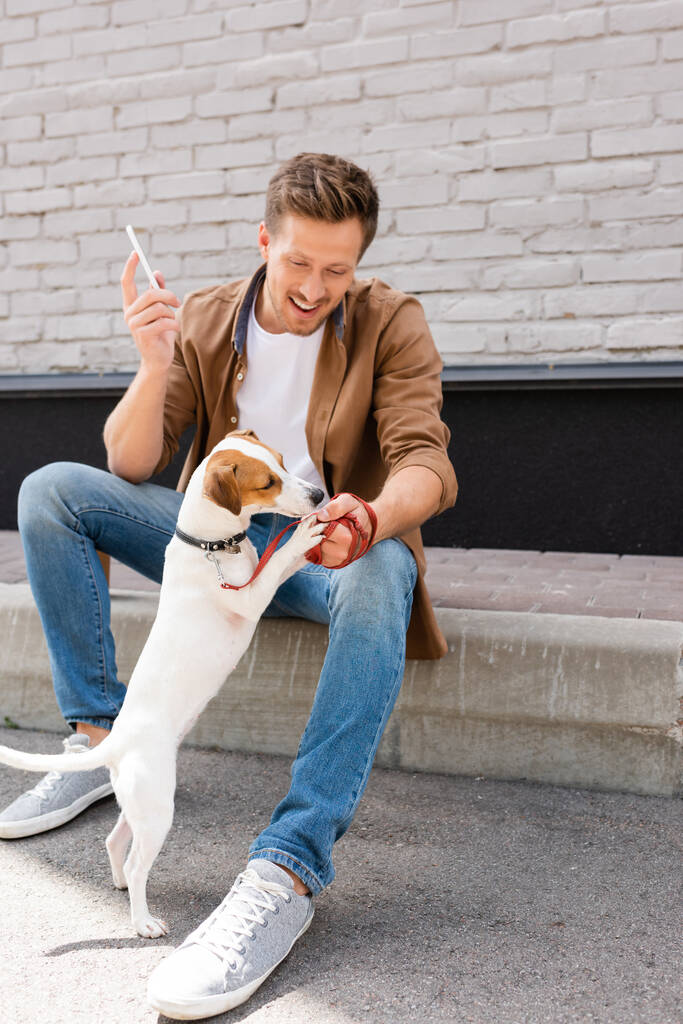 Jeune homme jouant avec jack russell terrier tout en tenant smartphone sur la rue urbaine  - Photo, image