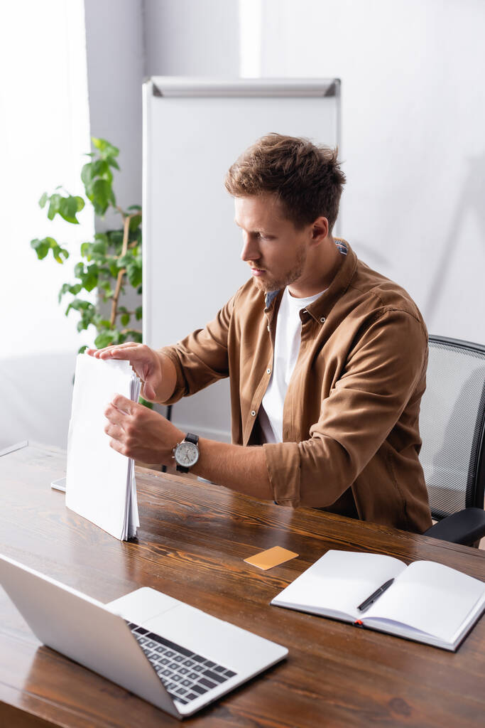 Concentration sélective de l'homme d'affaires tenant des papiers près de portable et ordinateur portable dans le bureau  - Photo, image