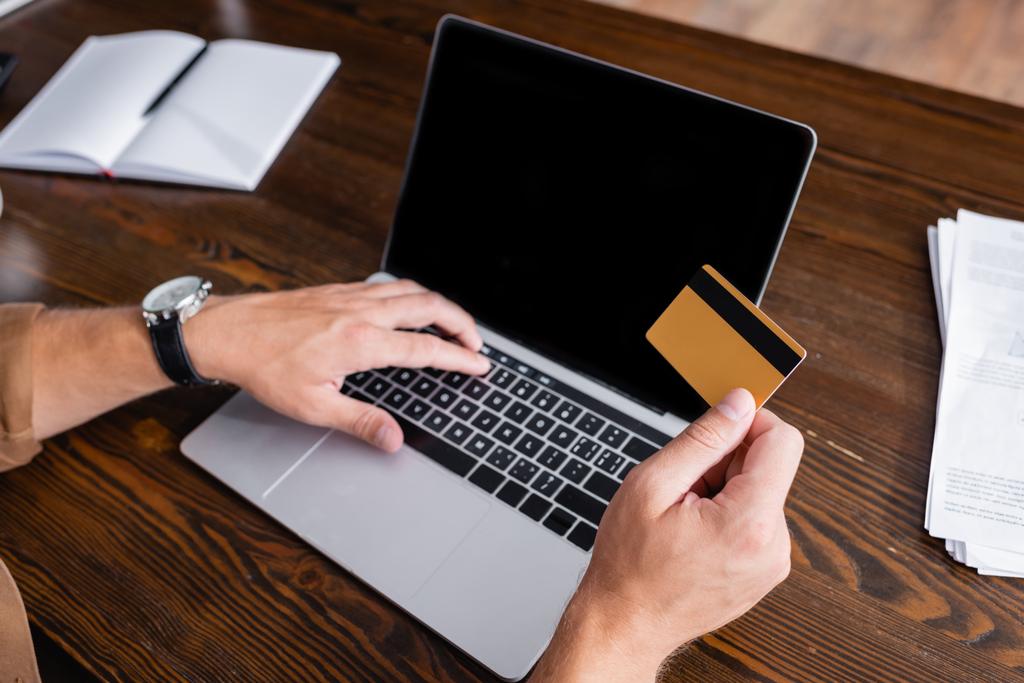 Vista recortada del hombre de negocios con tarjeta de crédito y el uso de la computadora portátil cerca de los papeles en la oficina  - Foto, imagen