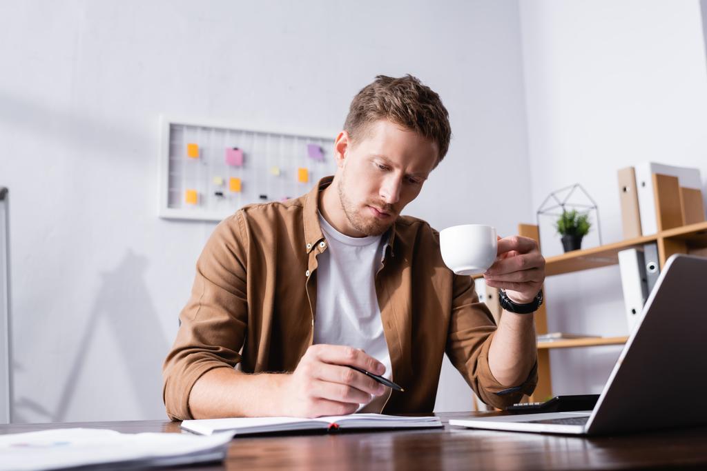 Selective focus of businessman writing on notebook and drinking coffee in office  - Photo, Image