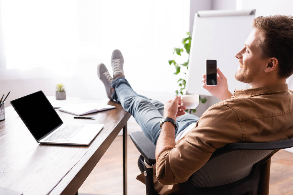 Вибірковий фокус молодого бізнесмена, який тримає чашку кави та мобільний телефон біля ноутбука на офісному столі
  - Фото, зображення