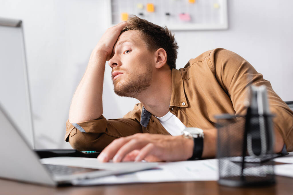 Вибірковий фокус втомленого бізнесмена з рукою біля голови за допомогою ноутбука під час роботи в офісі
  - Фото, зображення